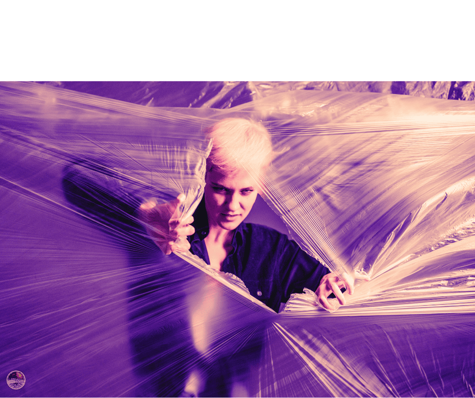 A person with short light-colored hair is partially wrapped in clear plastic, creating a dramatic effect with purple lighting.