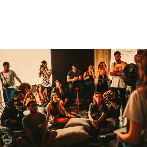 A group of people sitting and listening attentively while another person speaks or presents, surrounded by a casual setting with cushions and cameras.